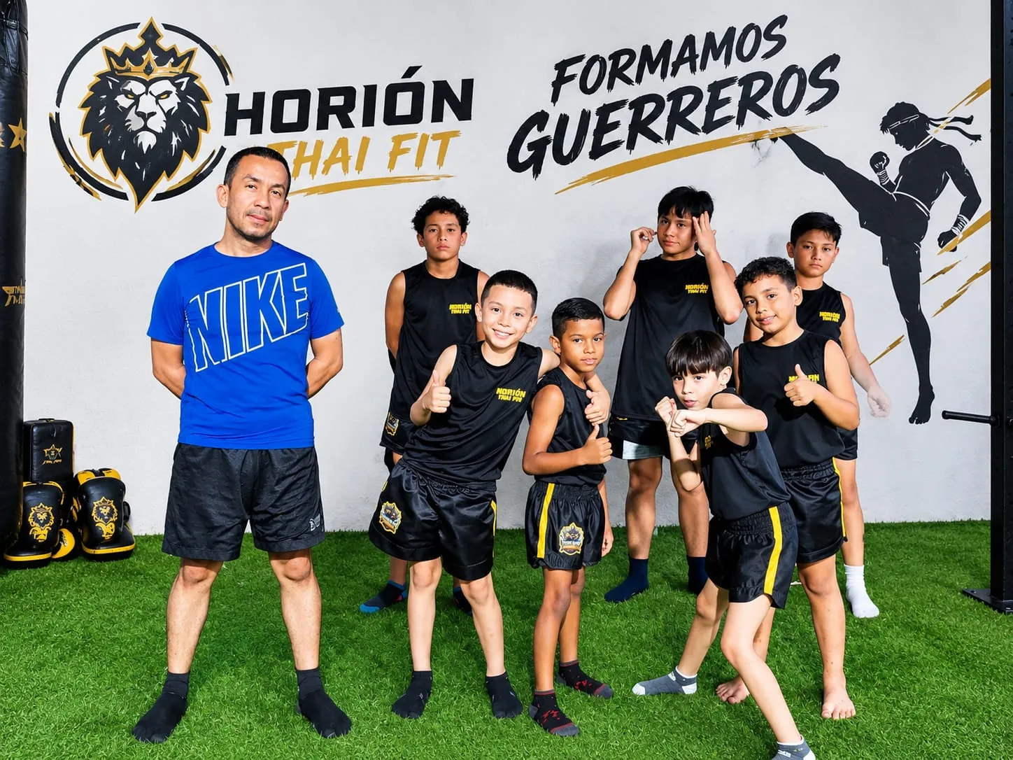 Un instructor con un grupo de niños con uniforme de Horión Thai Fit frente al muro con el logo del león